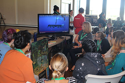 Girl Scout STEM participants crowd around the station showing the Wave Glider in action on a video screen